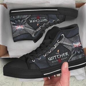 May include: A pair of black high-top sneakers featuring a graphic print of The Witcher Wild Hunt logo and a wolf medallion design. The shoes have black laces, black rubber toe caps, and white trim along the sole, presented inside a white box.