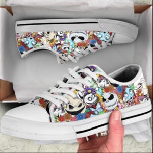 May include: White canvas sneakers with a colorful pattern featuring cartoon characters and floral designs. The shoes have white laces, a white rubber toe cap, and a black stripe along the sole. The sneakers are in a cardboard box.