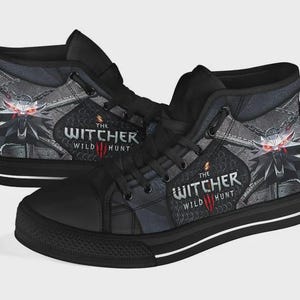 May include: A pair of black high-top sneakers featuring a graphic print of The Witcher Wild Hunt logo and a wolf medallion with glowing red eyes. The shoes have black laces, black eyelets, and a black rubber sole with a thin white horizontal stripe.