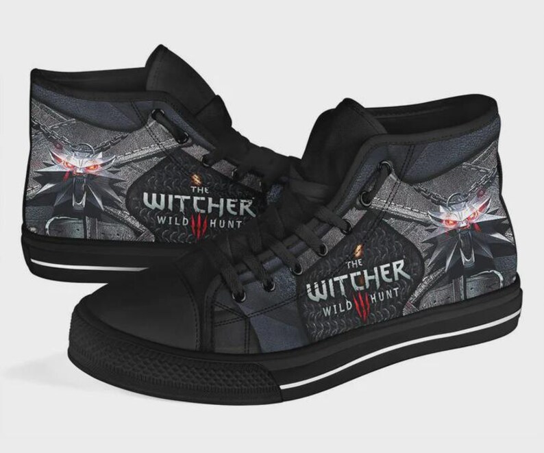 May include: A pair of black high-top sneakers featuring a graphic print of The Witcher Wild Hunt logo and a wolf medallion with glowing red eyes. The shoes have black laces, black eyelets, and a black rubber sole with a thin white horizontal stripe.