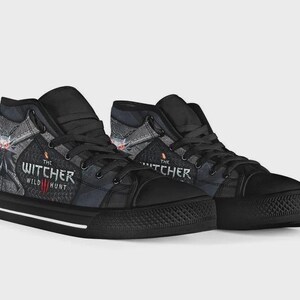 May include: A pair of black high-top sneakers featuring a graphic print from The Witcher 3 Wild Hunt. The shoes have black laces, black rubber toe caps, and a white stripe along the black sole. The side panels display the game logo and artwork.