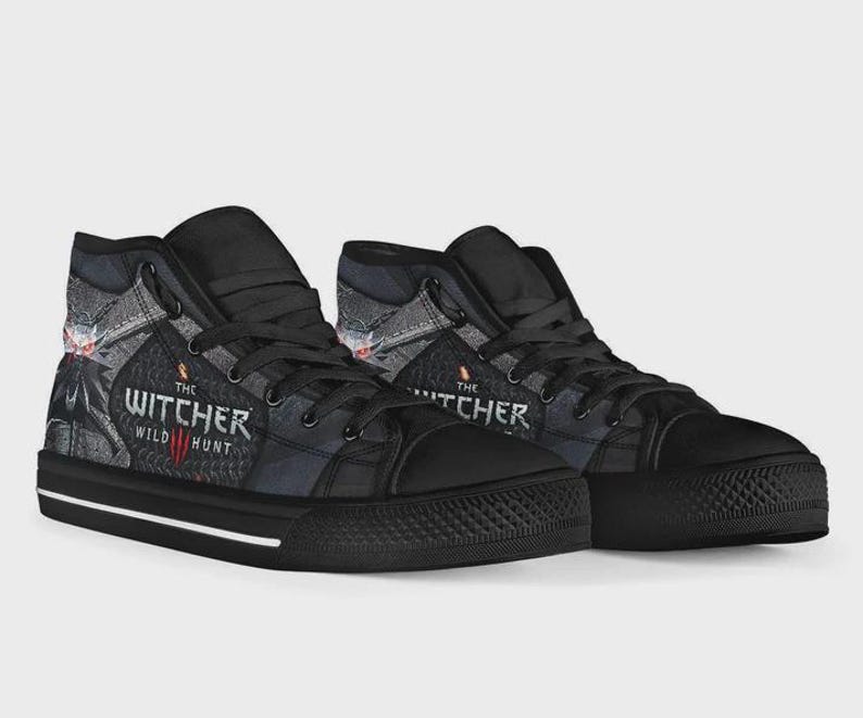 May include: A pair of black high-top sneakers featuring a graphic print from The Witcher 3 Wild Hunt. The shoes have black laces, black rubber toe caps, and a white stripe along the black sole. The side panels display the game logo and artwork.