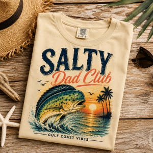 Pode incluir: Camiseta creme com o texto "SALTY Dad Club" em azul marinho e laranja. Um gr&aacute;fico de um peixe saltando para fora do oceano com um p&ocirc;r do sol ao fundo. As palavras "GULF COAST VIBES" s&atilde;o impressas na parte inferior.