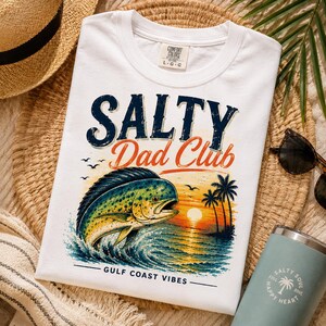 Pode incluir: Camiseta branca com o texto "SALTY Dad Club" em azul marinho e laranja. A camiseta apresenta um gr&aacute;fico colorido de um peixe saltando para fora do oceano com um fundo de p&ocirc;r do sol. O texto "GULF COAST VIBES" est&aacute; abaixo.