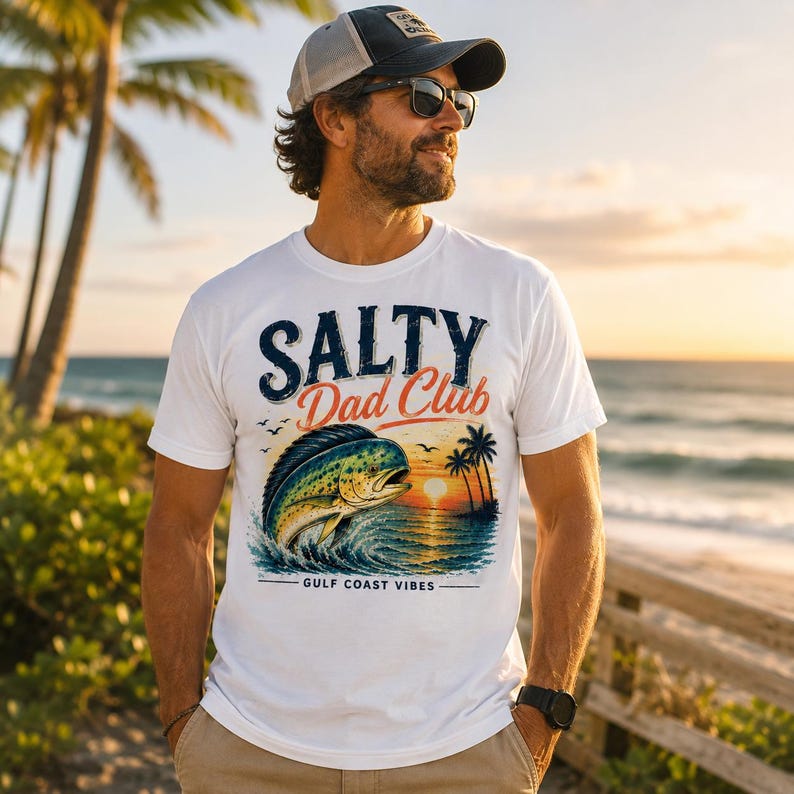 Pode incluir: Camiseta branca com o texto "Salty Dad Club" em azul marinho e laranja. A camisa apresenta um gr&aacute;fico de um peixe saltando para fora da &aacute;gua com um fundo de p&ocirc;r do sol. As palavras "Gulf Coast Vibes" s&atilde;o impressas abaixo.