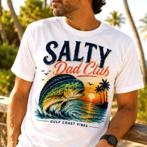 Pode incluir: Camiseta branca com um gr&aacute;fico colorido apresentando um peixe saltando das ondas do oceano ao p&ocirc;r do sol, palmeiras e o texto "SALTY Dad Club" e "GULF COAST VIBES".