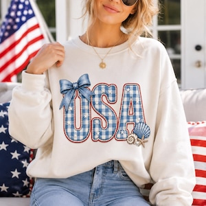 May include: Cream-coloured sweatshirt featuring the word "USA" in a blue and white gingham pattern, outlined in red. The design includes a blue bow and seashell graphics. An American flag is visible in the background.