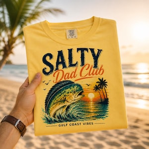 Pode incluir: Uma t-shirt amarela com as palavras "SALTY Dad Club" em azul marinho e vermelho. A t-shirt apresenta um gr&aacute;fico de um peixe a saltar para fora da &aacute;gua com um p&ocirc;r do sol e palmeiras. O texto "GULF COAST VIBES" est&aacute; por baixo do gr&aacute;fico.