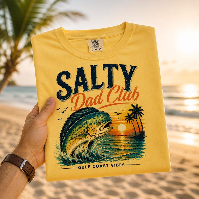 Pode incluir: Uma t-shirt amarela com as palavras "SALTY Dad Club" em azul marinho e vermelho. A t-shirt apresenta um gr&aacute;fico de um peixe a saltar para fora da &aacute;gua com um p&ocirc;r do sol e palmeiras. O texto "GULF COAST VIBES" est&aacute; por baixo do gr&aacute;fico.