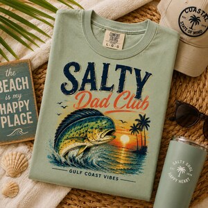 Pode incluir: Camiseta verde claro com o texto "Salty Dad Club" e um gr&aacute;fico de um peixe, palmeiras e um p&ocirc;r do sol. Um chap&eacute;u a condizer, &oacute;culos de sol e um copo tamb&eacute;m s&atilde;o vis&iacute;veis. Uma placa diz "A praia &eacute; o meu lugar feliz."