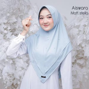 May include: A pale blue hijab with the text "Aiswara Matt. stella". The hijab features small decorative details. The woman is wearing a white long-sleeved shirt.