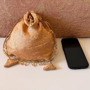 May include: A peach-colored drawstring pouch featuring a subtle floral pattern, gold-toned beaded fringe along the bottom edge, and two matching fabric tassels. A black smartphone is positioned next to the pouch for a visual size reference.