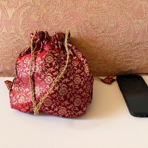 May include: A drawstring pouch made of red fabric with a gold floral pattern and gold braided cord handles. The pouch is positioned next to a black smartphone, providing a visual scale for the size of the bag in this product photograph.