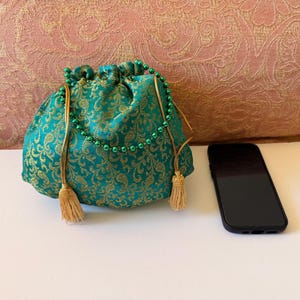 May include: A teal drawstring pouch featuring a gold floral pattern, a green beaded handle, and two gold tassels. The pouch is positioned next to a black smartphone to provide a sense of scale for the item's size in this visual presentation.