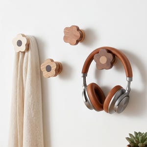 Op de afbeelding: Houten bloemvormige wandhaken in verschillende bruintinten. Eén haak houdt een beige sjaal vast, een andere een bruine koptelefoon. De haken zijn aan een witte muur bevestigd en bieden een decoratieve en functionele opbergoplossing.
