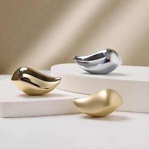 May include: Three bird-shaped figurines with smooth, rounded bodies. One figurine has a polished gold finish, one has a polished silver finish, and the third has a matte gold finish. Each piece features a curved, abstract form.