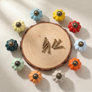 May include: A collection of colorful ceramic cabinet knobs in various shades including blue, yellow, red, orange, and green. Each knob has a flower-like shape with a brass-colored center. The knobs are arranged around a wooden slice with brass screws.