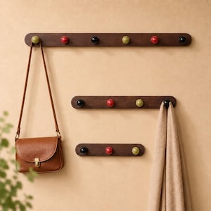 Op de afbeelding: Drie horizontale wandkapstokken van donkerbruin hout met afgeronde uiteinden, voorzien van bolvormige haken in rood, zwart en lichtgroen. Een bruine leren schoudertas hangt aan de bovenste kapstok en een beige stoffen sjaal hangt aan de middelste kapstok.