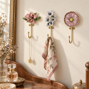 May include: Three wall-mounted hooks featuring decorative floral tops in pink, blue, and purple enamel. Each hook has a gold-toned metal finish and is secured with two screws. One hook holds a gold necklace, and another holds a floral patterned cloth.