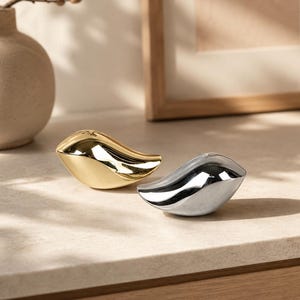 May include: Two small, smooth, bird-shaped figurines with a reflective finish. One figurine is polished gold, and the other is polished silver. Both objects feature a rounded, abstract form with a pointed tail and a smooth, curved body shape.