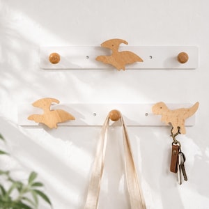 May include: Two white wooden wall-mounted coat racks with wooden dinosaur-shaped hooks. One rack has two dinosaur hooks and two wooden knobs. The other rack has one dinosaur hook, one wooden knob, and a dinosaur-shaped key holder.