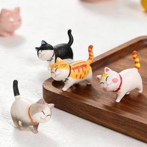 May include: A collection of small, smiling cat figurines in various colors. One is black with a white face, another is orange and yellow striped, and a third is white with a red collar. The figurines are on a wooden tray.