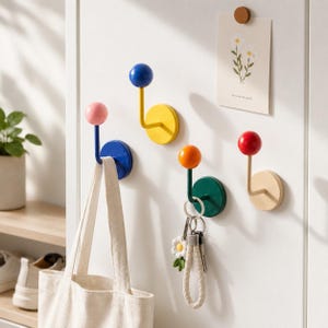 Op de afbeelding: Vier wandhaken met bolvormige uiteinden in roze, blauw, oranje en rood. Elke haak heeft een ronde basisplaat in bijpassende of contrasterende kleuren. Een witte canvas tas hangt aan de blauwe haak en een sleutelhanger hangt aan de groene haak.