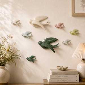 May include: A collection of nine ceramic bird wall decorations in various sizes and colors, including cream, light blue, pink, green, and light green. The central dark green bird features the name Charlotte engraved on its wing with a small heart.
