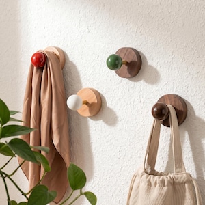 May include: A set of wall-mounted coat hooks with wooden bases and coloured knobs. One hook holds a tan scarf, another a beige drawstring bag. The knobs are red, green, white, and brown.