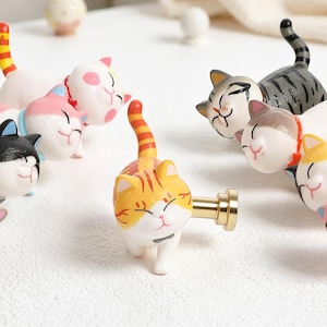 May include: A collection of colorful cat-shaped cabinet knobs. The knobs feature various designs, including orange tabby, gray striped, and white with pink spots. Each knob has a gold-colored metal base for easy installation.