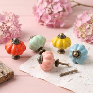 May include: A collection of colorful ceramic cabinet knobs in shades of orange, green, yellow, pink, and blue. Each knob has a unique pumpkin-like shape with a bronze-colored base and hardware. The knobs are arranged on a textured paper surface.