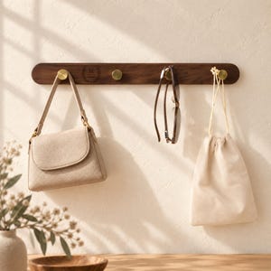 Può includere: Un appendiabiti da parete orizzontale in legno scuro con quattro ganci circolari in ottone. Una borsa beige, un paio di occhiali da sole marroni e una pochette color crema con coulisse sono appesi ai ganci. Il legno presenta una lettera M incisa.