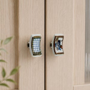 May include: Two decorative cabinet knobs. One knob features a blue and white grid pattern, and the other has a floral design with blue and orange accents. Both knobs have a white base with a gold trim and are mounted on a light wood cabinet.