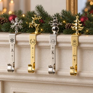 May include: Four metal stocking holders with flat vertical shafts and curved hooks. The first is silver with a snowflake topper and letter A. The second is gold with a reindeer topper, initials JM, and stars. The third is silver with a snowman topper, a hat icon, and letter K. The fourth is gold with a tree topper, initials LK, and the year 2026.