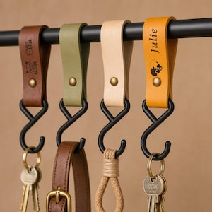 May include: Four leather loop hangers in brown, olive green, beige, and mustard yellow, each secured with a brass rivet and hanging from a black metal S-hook on a black rod. The brown loop features the name Ella, and the mustard loop features the name Julie.
