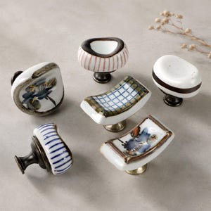 May include: A collection of six ceramic cabinet knobs with various designs. The knobs feature hand-painted patterns, including floral motifs, stripes, and grid patterns. Each knob has a unique shape and is mounted on a metal base, offering a decorative touch for furniture.