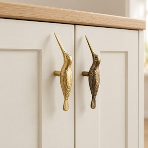 May include: Two metallic gold-toned cabinet knobs shaped like hummingbirds with detailed wing and feather textures. The knobs are mounted vertically on two adjacent off-white cabinet doors with a light wood countertop visible at the top edge.