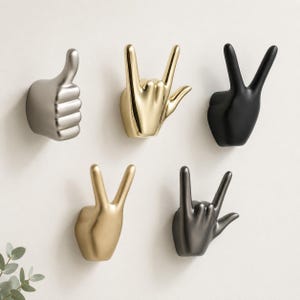 May include: A set of five wall hooks shaped like hands in various gestures. The collection includes a silver thumbs-up, a gold I-love-you sign, a matte black peace sign, a gold peace sign, and a dark metallic grey I-love-you sign.