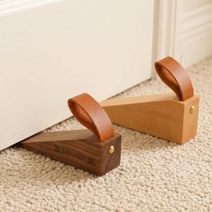 May include: Two wooden doorstops with leather handles. One is a dark brown color, and the other is a light brown color. The dark brown doorstop has the word "JAPANESE" printed on it. Both doorstops have a brass-colored stud.