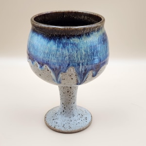 May include: A ceramic goblet featuring a speckled light gray base and a stem that transitions into a deep blue and turquoise glaze at the rim. The glaze drips downward in a vertical pattern over the speckled surface, creating a distinct contrast.
