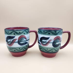 May include: Two ceramic mugs with a red base and handle, and a green and white patterned design. The mugs have a dark red rim and a decorative floral pattern in red and dark blue.