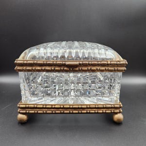 May include: A rectangular clear glass box with a faceted diamond-cut pattern on the lid and sides. The box is encased in a metallic bronze-toned frame with a beaded texture and is supported by four small, rounded ball-shaped feet at the base.