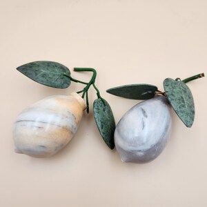 May include: Two decorative fruit sculptures shaped like lemons. One is crafted from cream and tan marbled stone, the other from grey and white marbled stone. Each features dark green, textured, pointed leaves attached to a thin green stem.