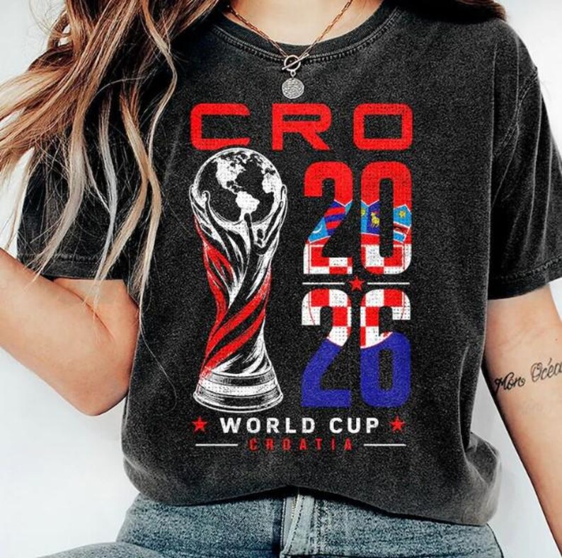 May include: A charcoal grey t-shirt featuring a graphic print with the text CRO 2026 and WORLD CUP CROATIA. The design includes a stylised illustration of a trophy topped with a globe, and the numbers 2026 filled with a red and white checkerboard pattern.