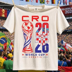 May include: An off-white short-sleeve t-shirt featuring a graphic print with the text CRO 2026 WORLD CUP CROATIA. The design includes a stylised trophy illustration and the numbers 2026 filled with a red and white chequered pattern.