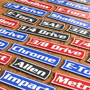 May include: A collection of rectangular tool box labels in red, blue, black, and white. The labels feature text including 1/4 Drive, 3/8 Drive, E-Torx, Shallow, SAE, Chrome, Allen, Impact, and Metric, designed for organizing various mechanical tools.