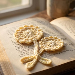May include: A handmade, crochet flower bookmark in soft yellow, resting on the pages of an open book. The bookmark features two flower heads and a stem, perfect for book lovers. The book is open to a page with printed text.