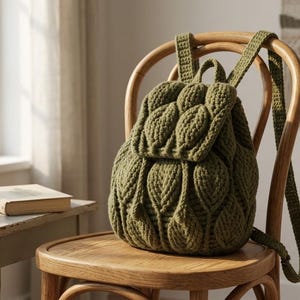 Op de afbeelding: Olijfgroene gebreide rugzak met een bladerpatroon, op een houten stoel. De tas heeft verstelbare bandjes en een klepsluiting. Een kleine vetplant in een terracotta pot, een mok en een boek staan op een bijzettafel.