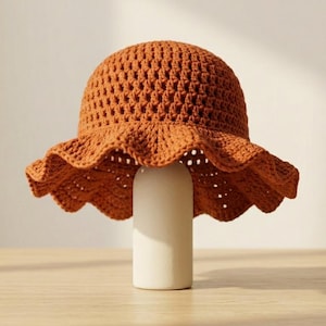 Puede incluir: Un sombrero de pescador de ganchillo color óxido con un patrón de punto abierto texturizado y un ala ancha, ondulada y festoneada. El sombrero presenta una copa redondeada y está hecho de hilo, mostrando un tono naranja-marrón profundo.