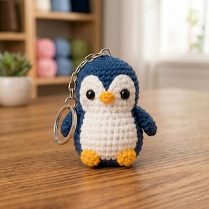 May include: A handmade crochet penguin keyring. The penguin has a blue body, white belly, and orange beak and feet. It features black safety eyes and a silver keyring. The penguin is approximately 7.6 cm tall.